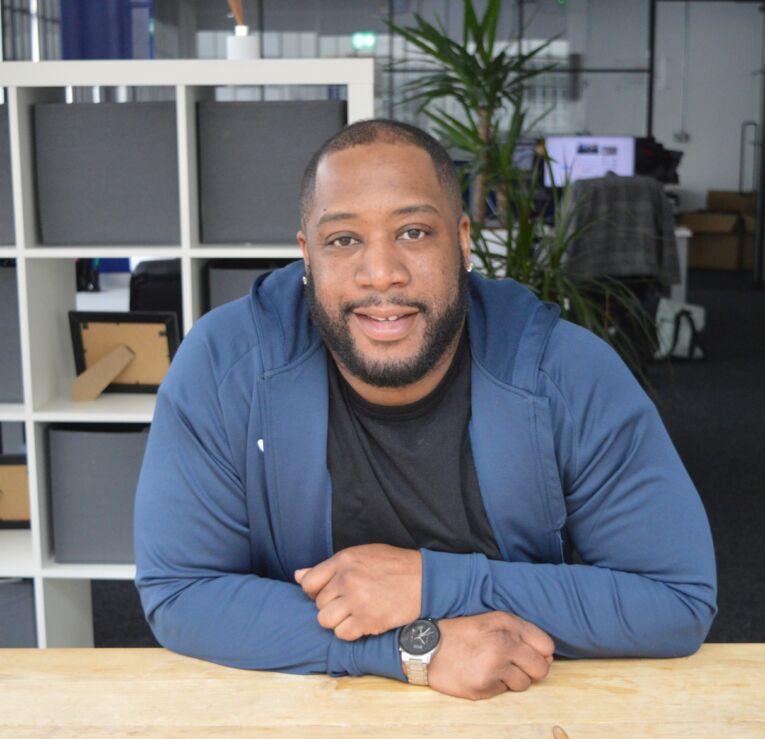 A picture of a Black Caribbean male smiling, sat down, resting arms on a wooden table.